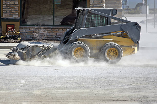 Small multi purpose construction vehicle using a power sweeper attachment cleans a restaurant parking lot of sand and debris.